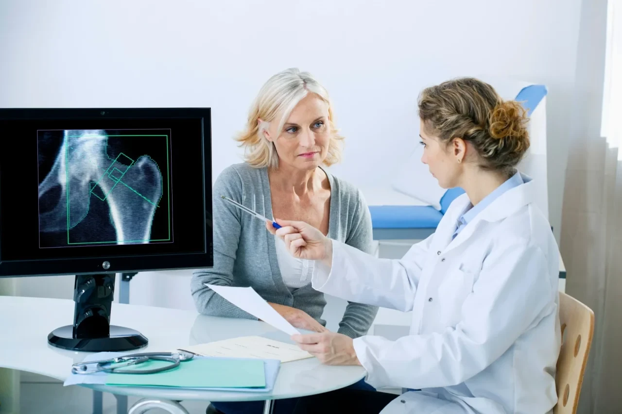 Patientin und Ärztin sitzen am Tisch mit einem Bildschirm, der eine Knochenaufnahme zeigt