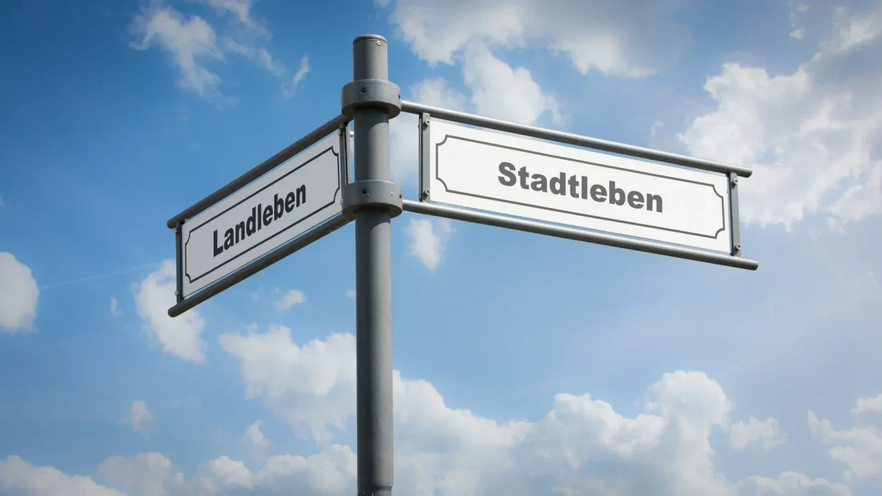 Ein Wegweiser mit zwei Schildern mit den Aufschriften „Landleben“ und „Stadtleben“ an einer Metallstange vor blauem Himmel mit Wolken.