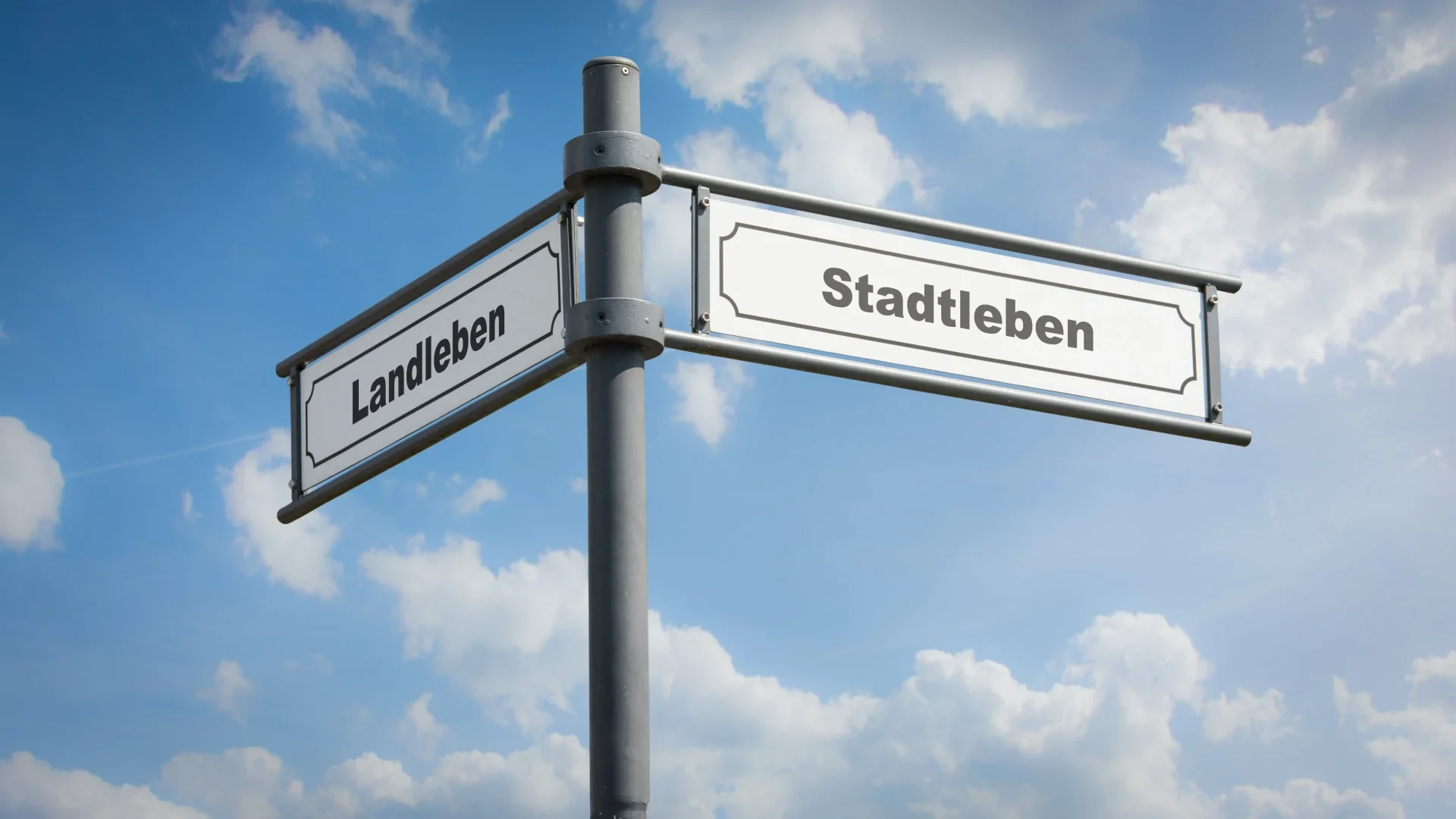 Ein Wegweiser mit zwei Schildern mit den Aufschriften „Landleben“ und „Stadtleben“ an einer Metallstange vor blauem Himmel mit Wolken.