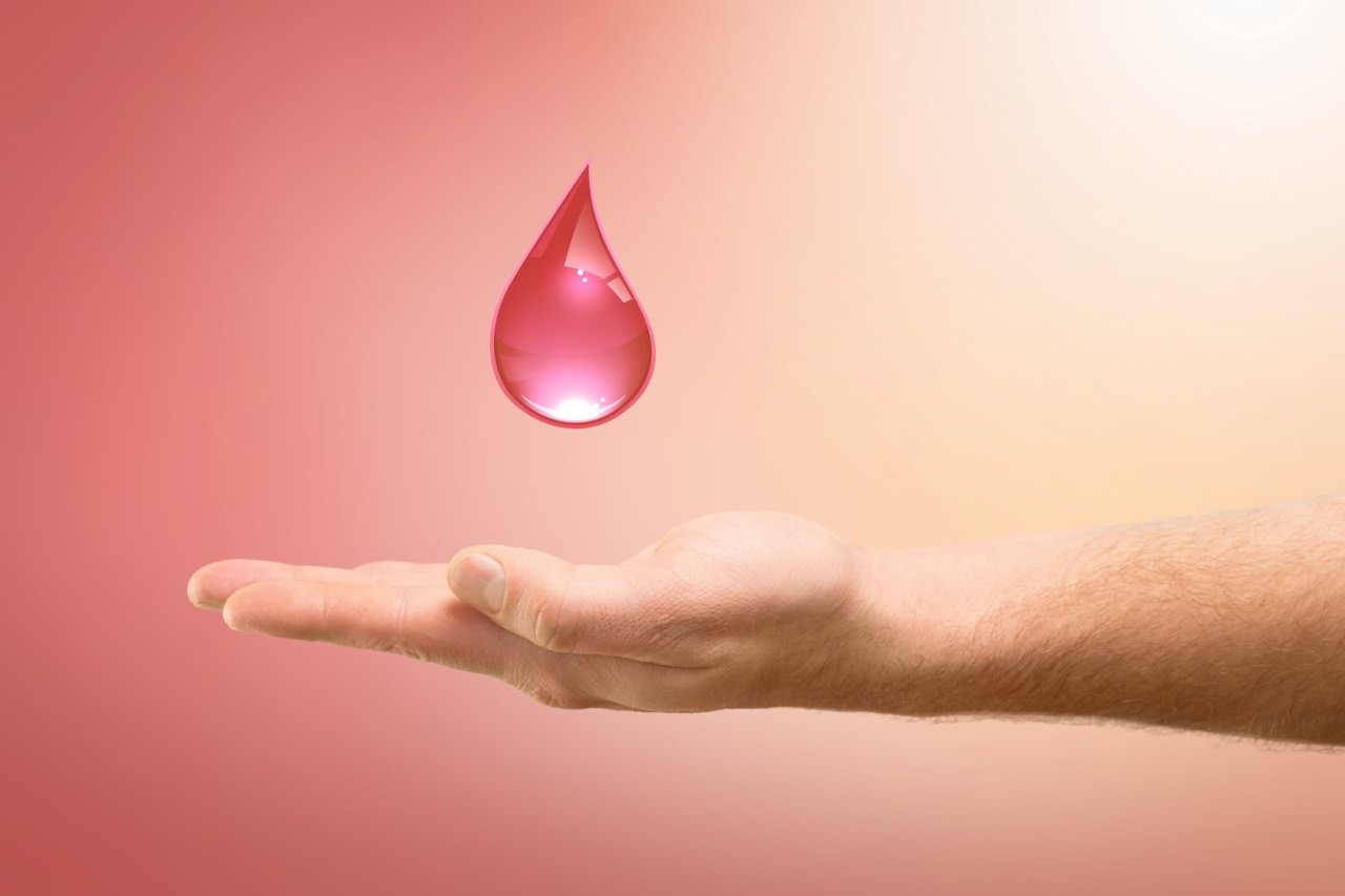 Eine ausgestreckte Hand hält einen roten Blutstropfen vor einem sanften rosa Hintergrund