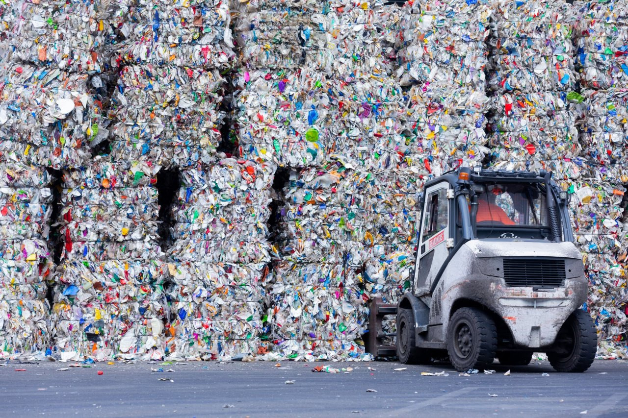 Ein kleines Arbeitsfahrzeug steht vor meterhohen, dicht gepressten Ballen aus buntem Plastikmüll auf einem Recyclingplatz