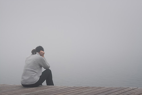Eine Person sitzt auf einem Holzsteg am Wasser, umgeben von dichtem Nebel. Die Haltung wirkt nachdenklich und zurückgezogen