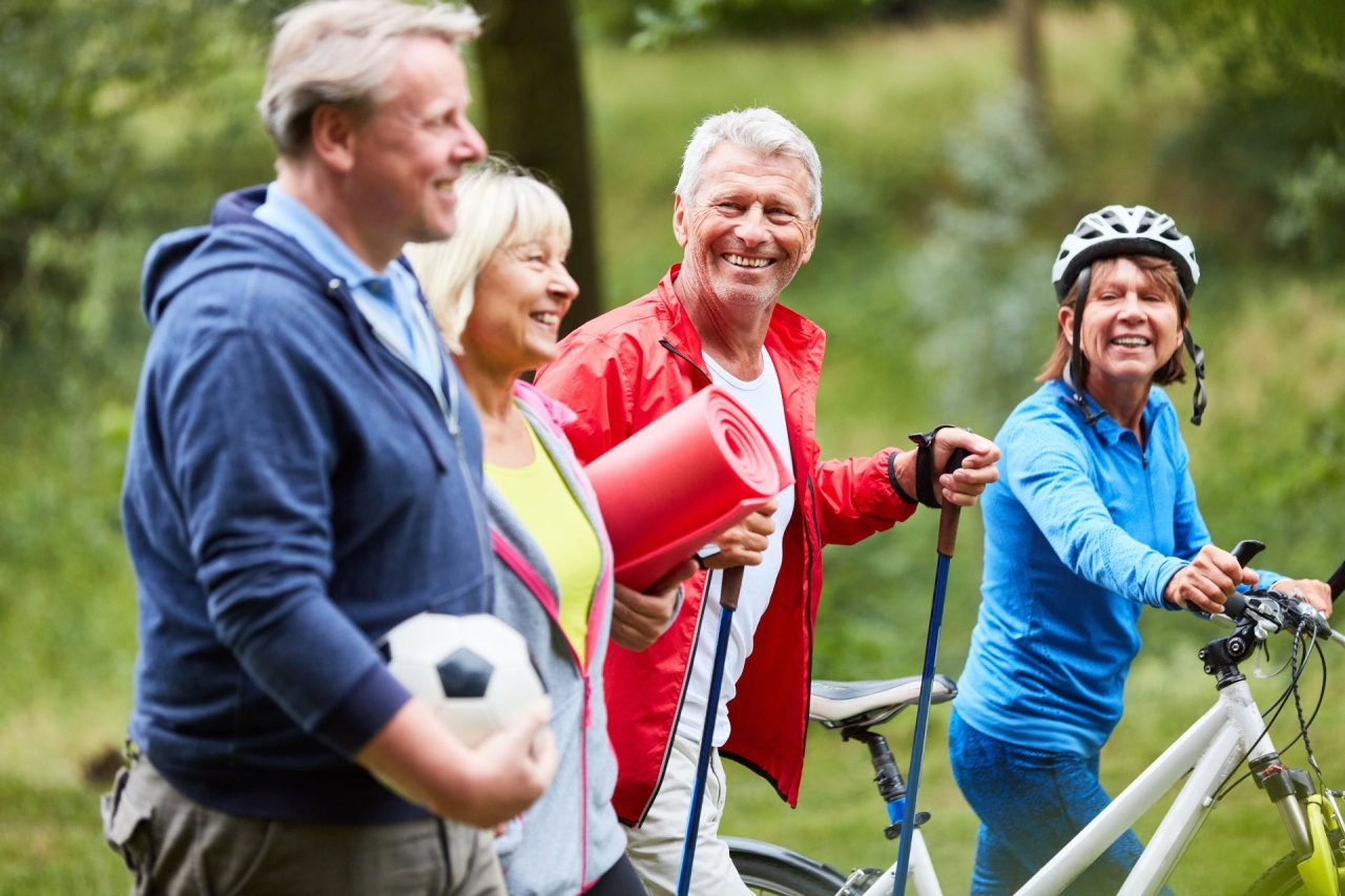 Eine Gruppe erwachsener Menschen ist gemeinsam draußen unterwegs, einige mit Wanderstöcken, eine Person mit Fahrrad; sie bewegen sich in einer grünen, natürlichen Umgebung und wirken aktiv und gut gelaunt.