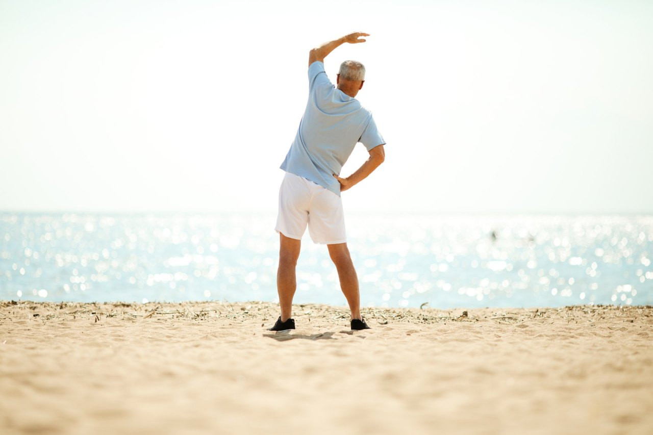 Ein älterer Herr in Sportkleidung steht am Strand und führt eine seitliche Dehnübung aus, mit Meer und hellem Himmel im Hintergrund.