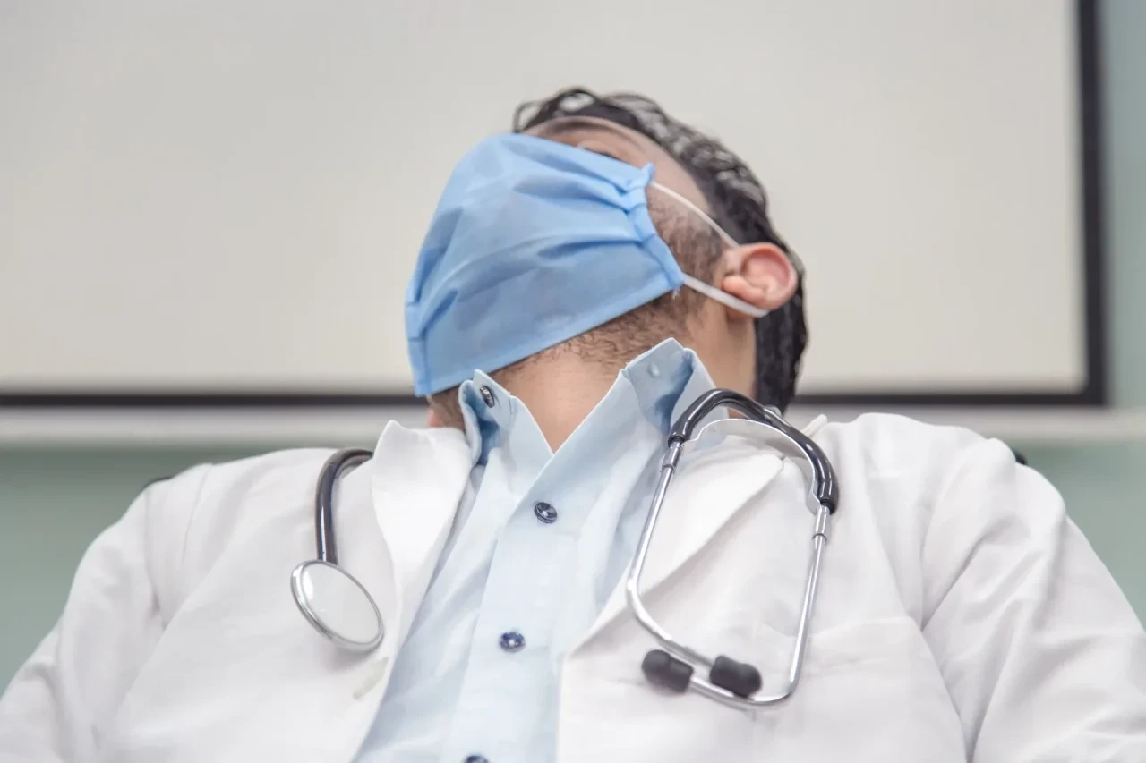 Ein müder, erschöpfter Arzt schläft an seinem Schreibtisch im Krankenhaus oder in der Praxis. Er trägt Maske und ein Stethoskop um den Hals.