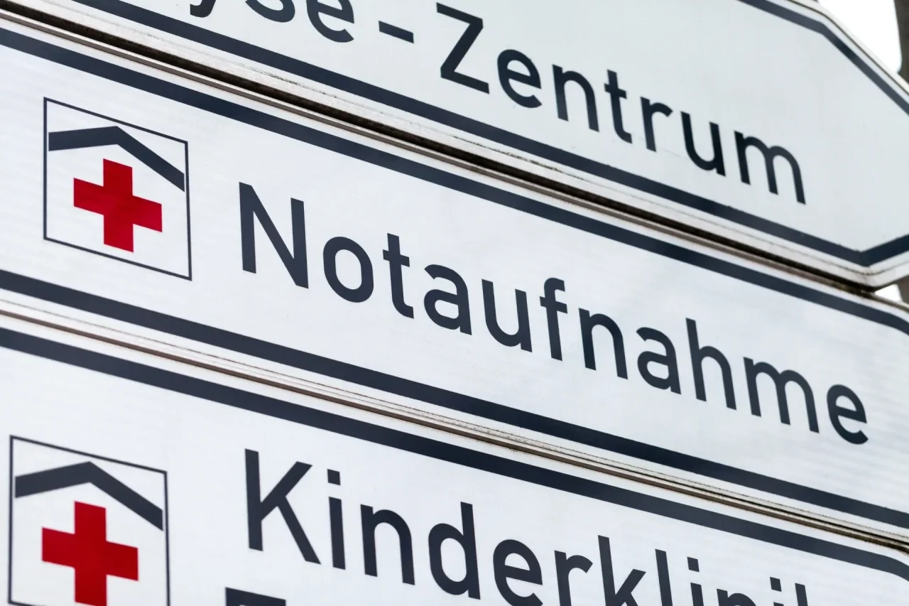 Nahaufnahme eines weißen Wegweisers mit rotem Kreuz und der Aufschrift „Notaufnahme“ sowie weiteren Hinweisen zu Klinikbereichen.