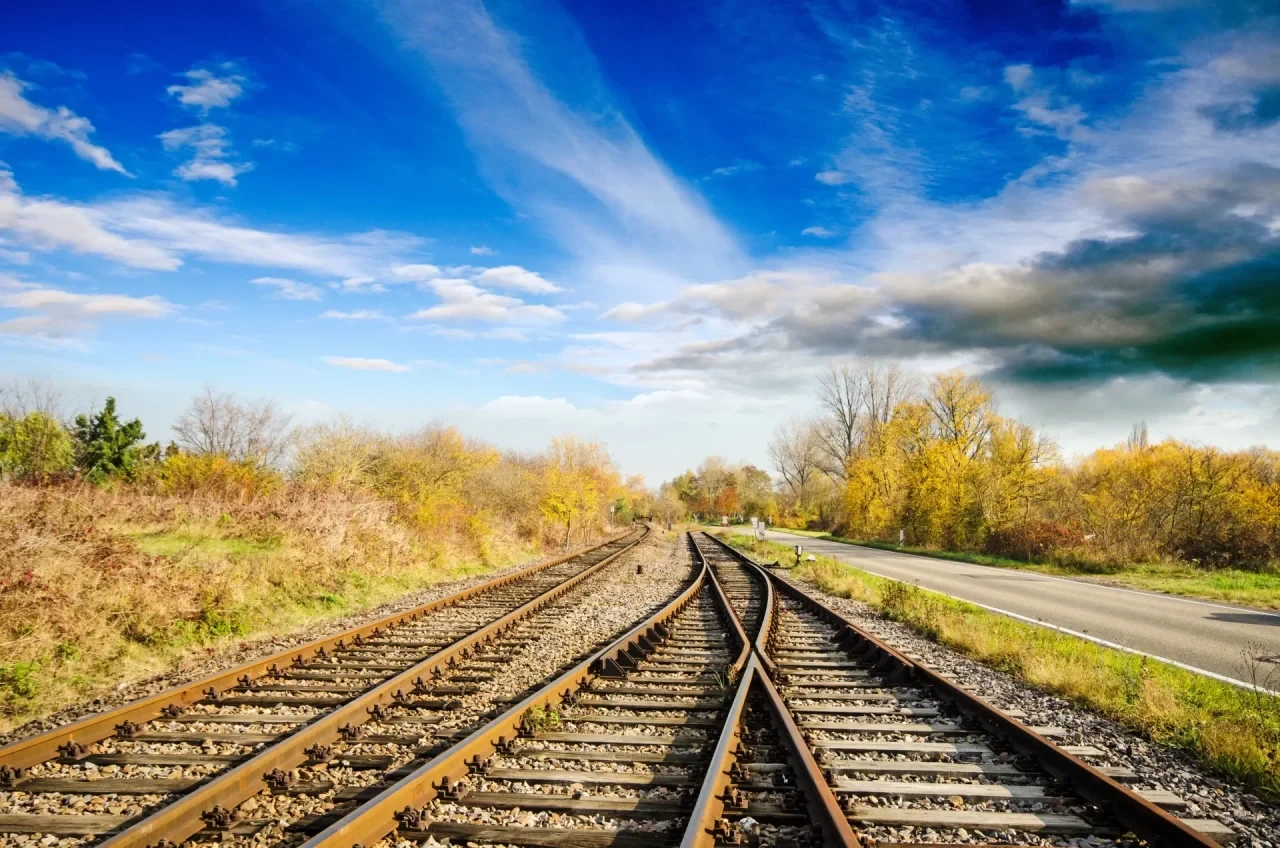 Zwei parallel verlaufende und sich teilende Eisenbahnschienen in einer ländlichen Landschaft mit Bäumen, Wiesen und blauem Himmel.