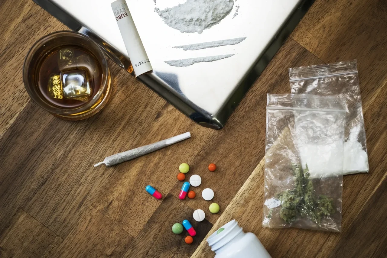 Whiskyglas, Tabletten in unterschiedlichen Farben, Cannabisblüten und weißes Pulver in einem Plastikbeutel sowie eine aufgerollte Banknote auf einem Holztisch – Symbolbild für Mischkonsum von Alkohol, Medikamenten und illegalen Drogen.