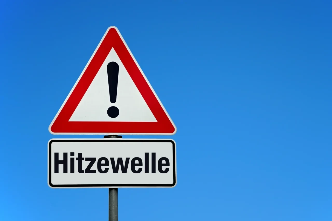 Rotes Warnschild mit Ausrufezeichen und Aufschrift „Hitzewelle" vor blauem Himmel.
