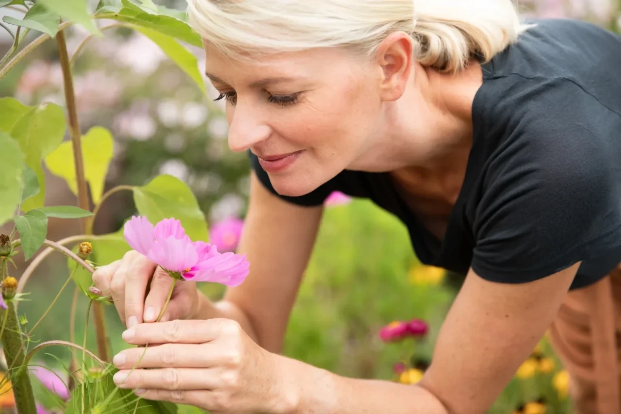 Eine Frau beugt sich in einem Garten zu einer Pflanze mit rosa Blüten, betrachtet diese aus nächster Nähe und nimmt ihren Duft wahr.
