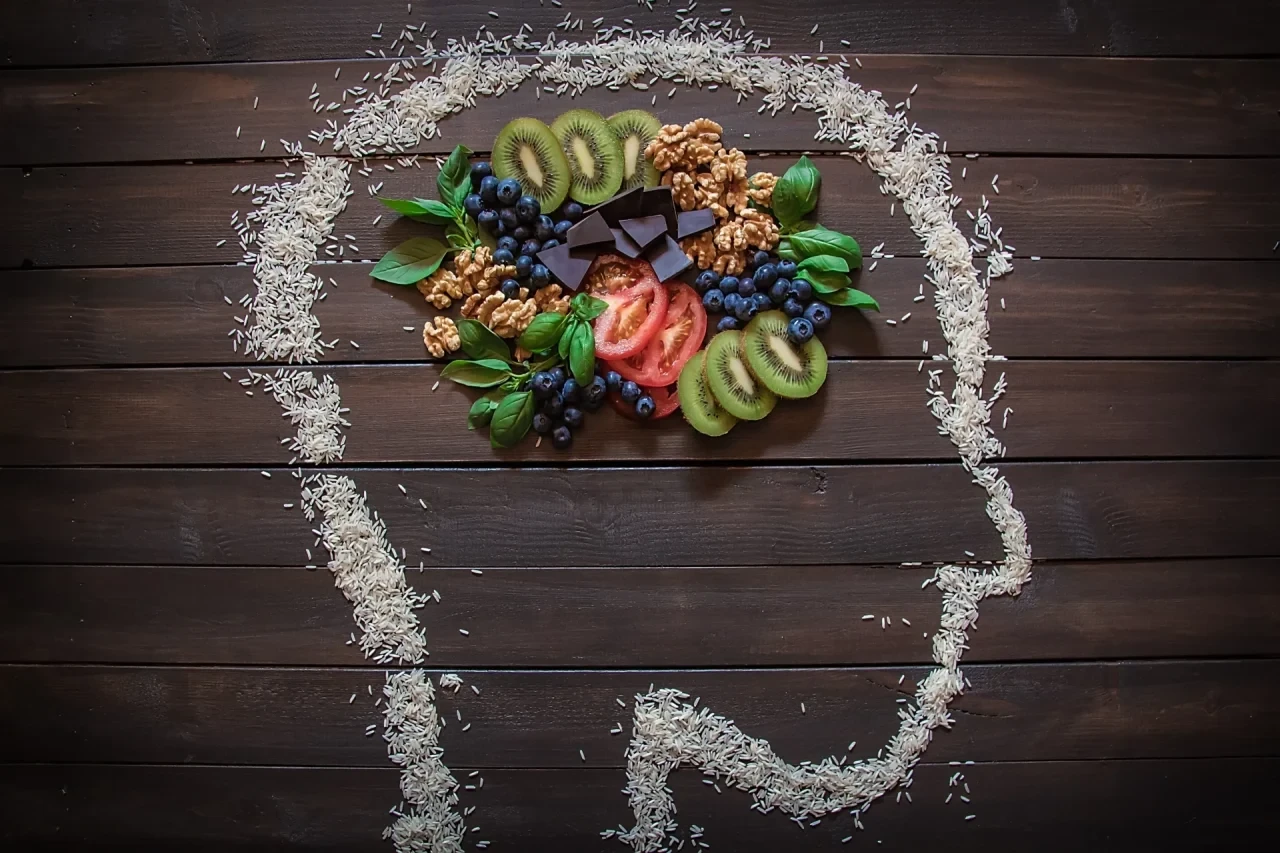 Aus Reiskörnern gestreute Silhouette eines menschlichen Kopfes im Profil auf dunklem Holz. Der Gehirnbereich ist mit einer bunten Auswahl an Lebensmitteln wie Kiwi, Beeren, Tomaten und Nüssen ausgelegt.