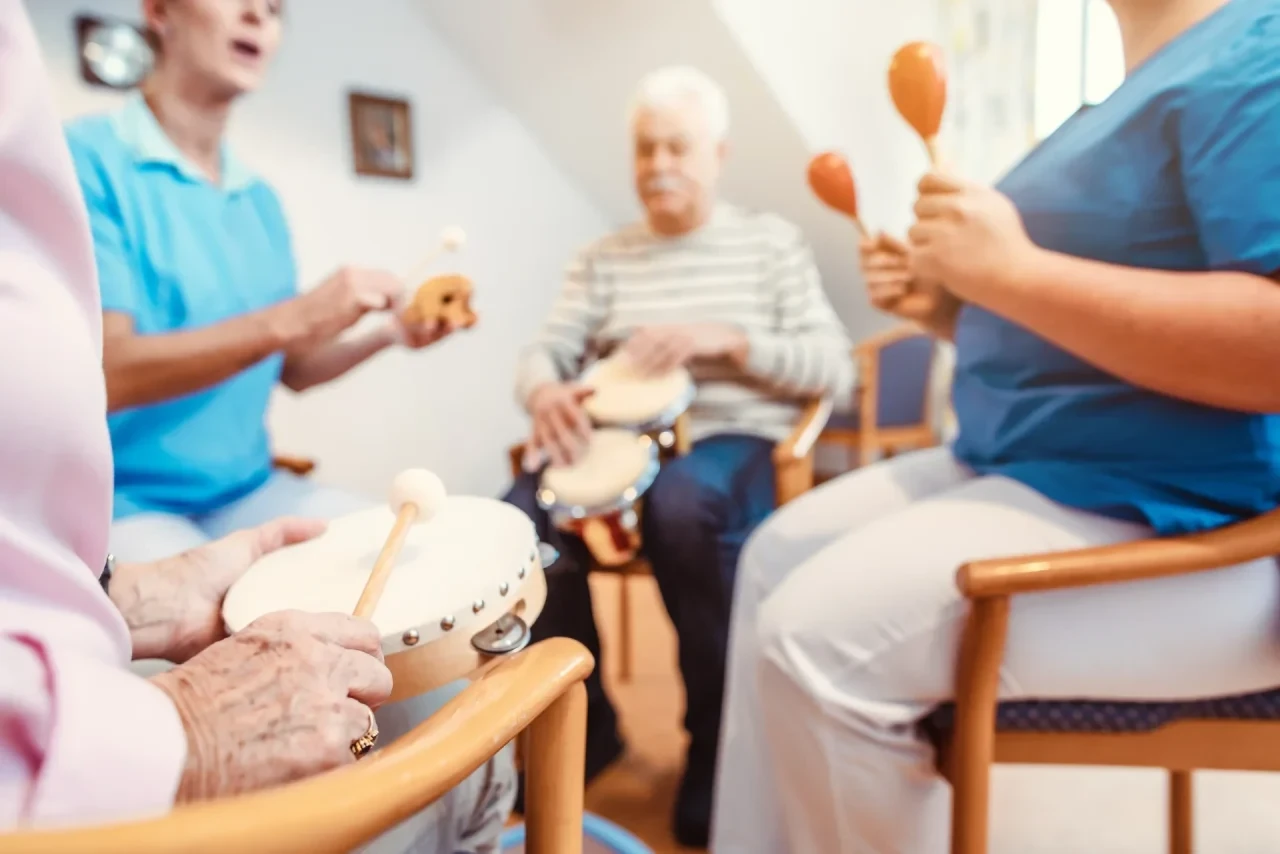 Senioren und Pflegekräfte sitzen im Kreis und musizieren gemeinsam mit verschiedenen Rhythmusinstrumenten wie Trommeln und Tamburinen in einem hellen Gemeinschaftsraum eines Pflegeheims.