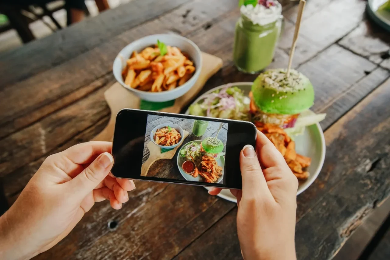 Eine Person hält ein Smartphone mit beiden Händen über einen Holztisch und fotografiert eine Mahlzeit bestehend aus Nudeln, einem grünen Burger mit Pommes und einem Smoothie, um die Kalorien per App zu erfassen.