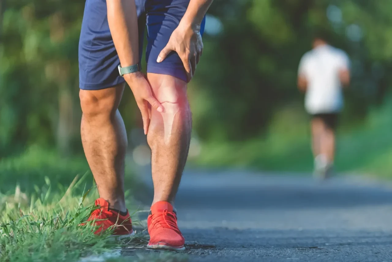 Nahaufnahme eines Sportlers in Laufkleidung im Park, der sich mit beiden Händen an sein schmerzendes Knie fasst. Das Knie ist durch eine rote Färbung als Schmerzzentrum markiert, zudem sind die Ober- und Unterschenkelknochen angedeutet. Im Hintergrund ist unscharf ein weiterer Läufer zu sehen.