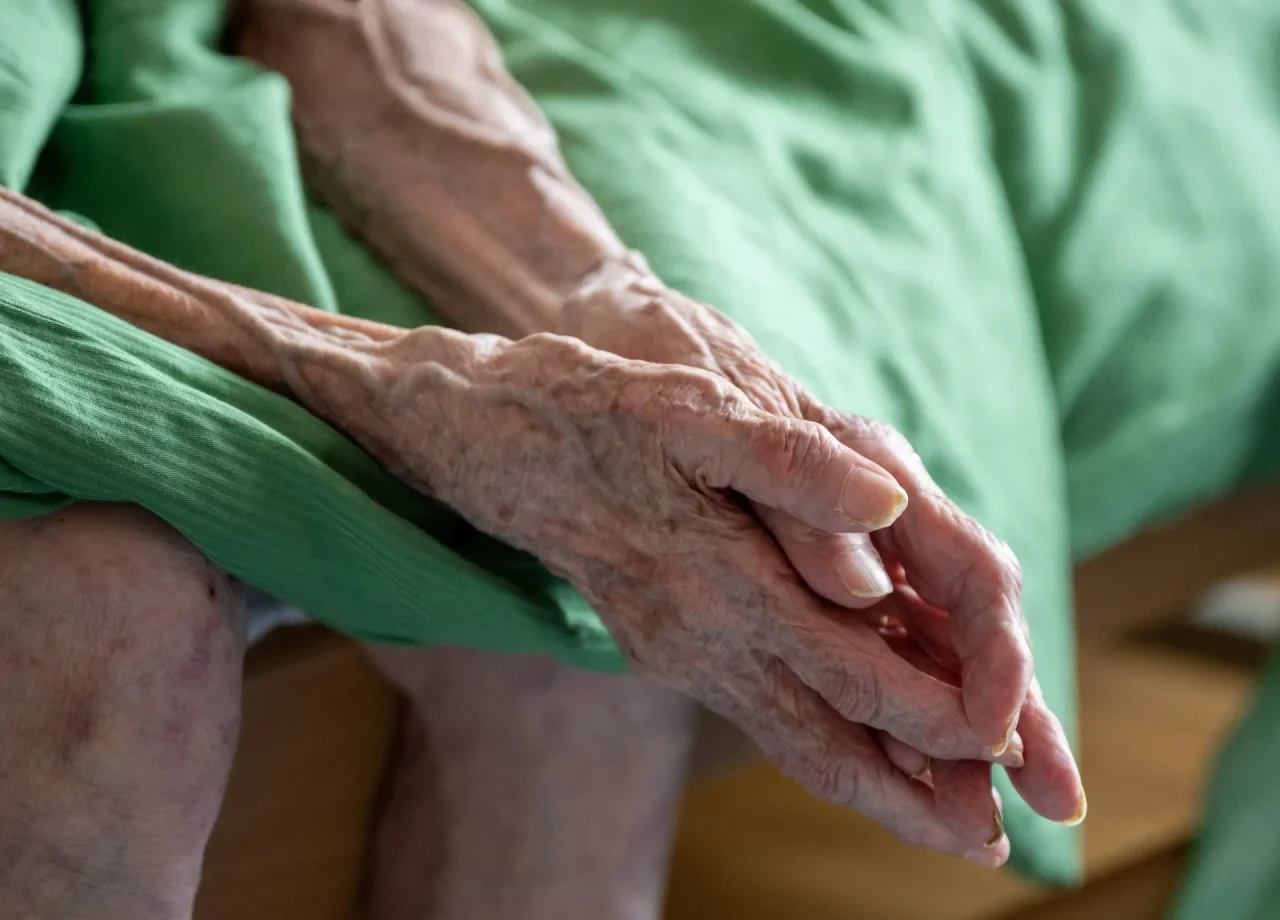 Eine pflegebedürftige Seniorin sitzt auf ihrem Bett, aufgenommen beim Besuch einer ambulanten Pflegekraft in der Wohnung der Patientin.