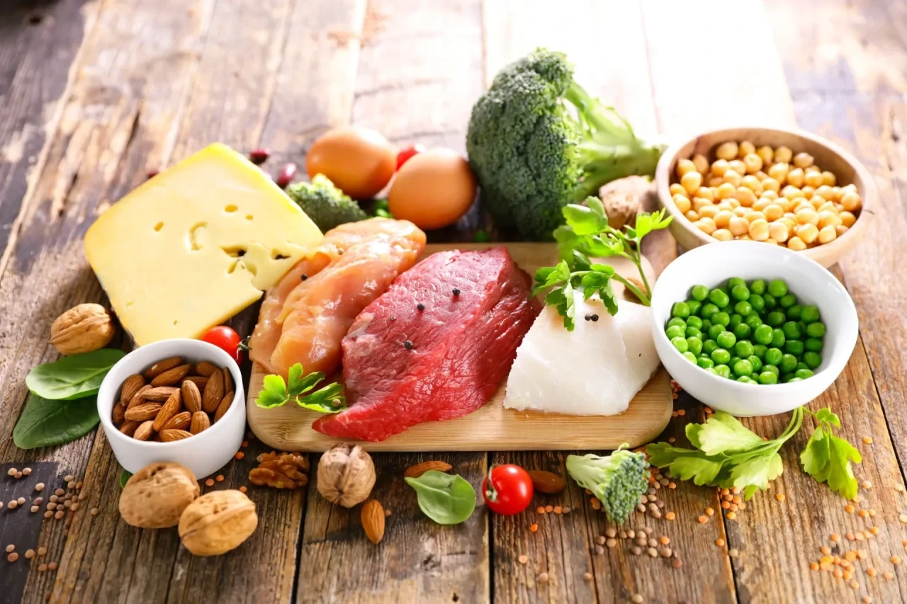 Auswahl eiweißreicher Lebensmittel mit rohem Fleisch, Fisch, Käse, Eiern, Hülsenfrüchten, Nüssen und frischem Gemüse auf einem Holztisch