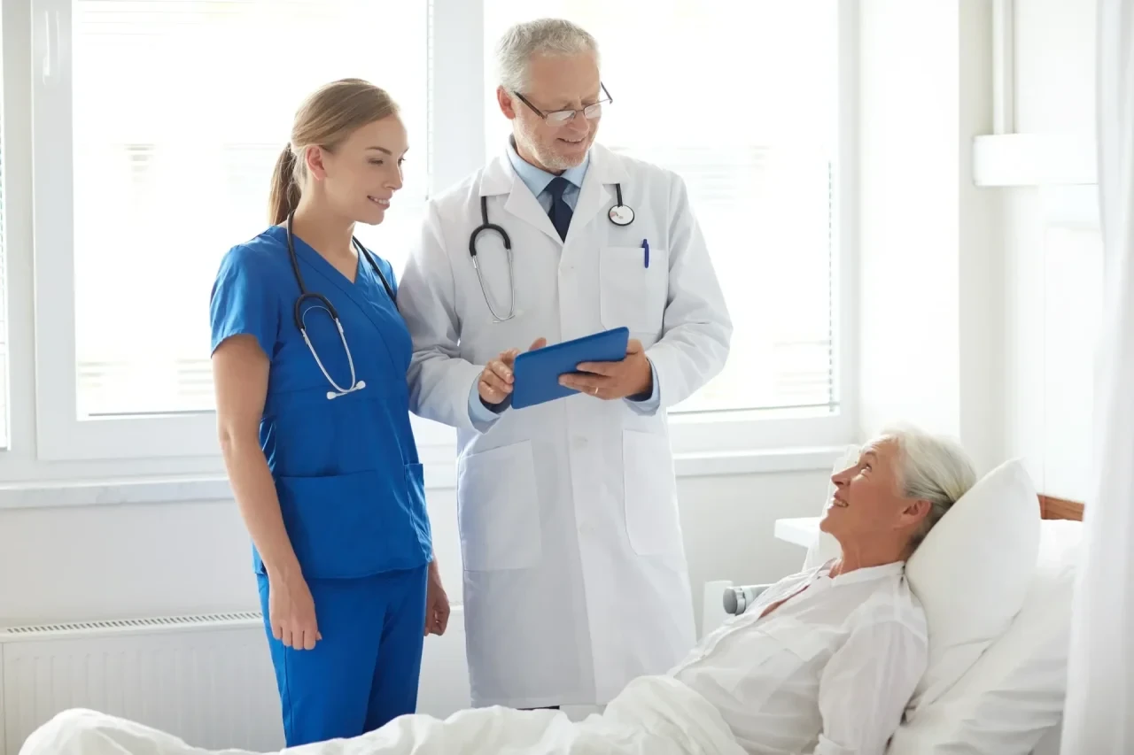 Eine ältere Patientin liegt im Krankenhausbett, ein Arzt und eine Pflegekraft stehen daneben und besprechen Befunde auf einem Klemmbrett.