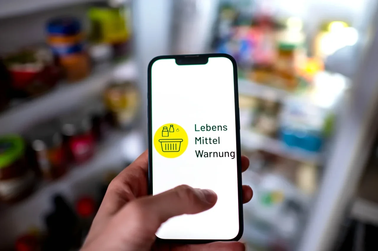 Die App «Lebensmittelwarnung» ist auf einem Smartphone vor einem geöffneten Kühlschrank zu sehen.