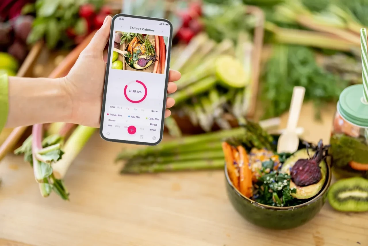 Hand hält Smartphone mit geöffneter Ernährungstracking-App über einer Schale mit frischem Gemüsesalat und buntem Gemüse auf einem Holztisch.