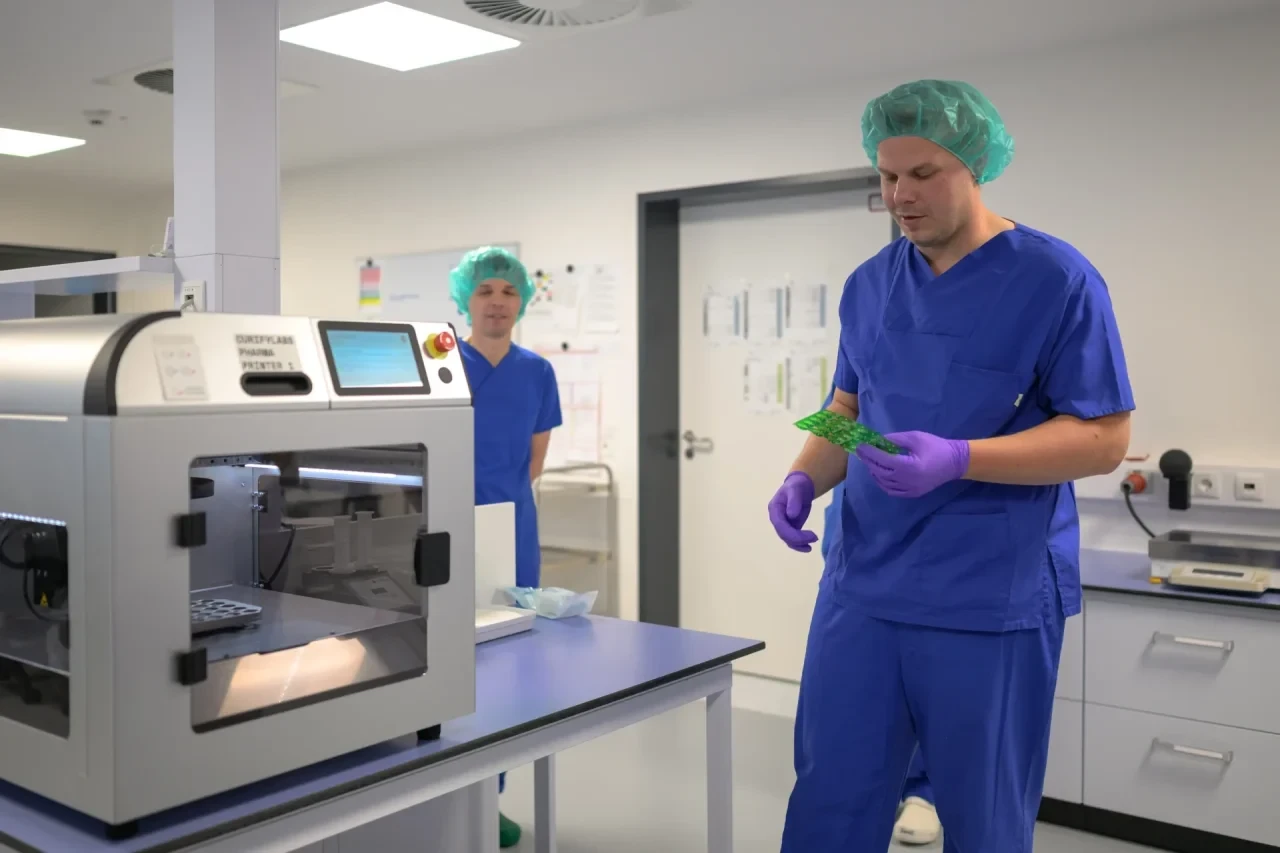 Renke Eggers (v. r.) geht mit einem Blister zum 3D-Arzneimitteldrucker, um ein Medikament in gewünschter Dosierung zu drucken.