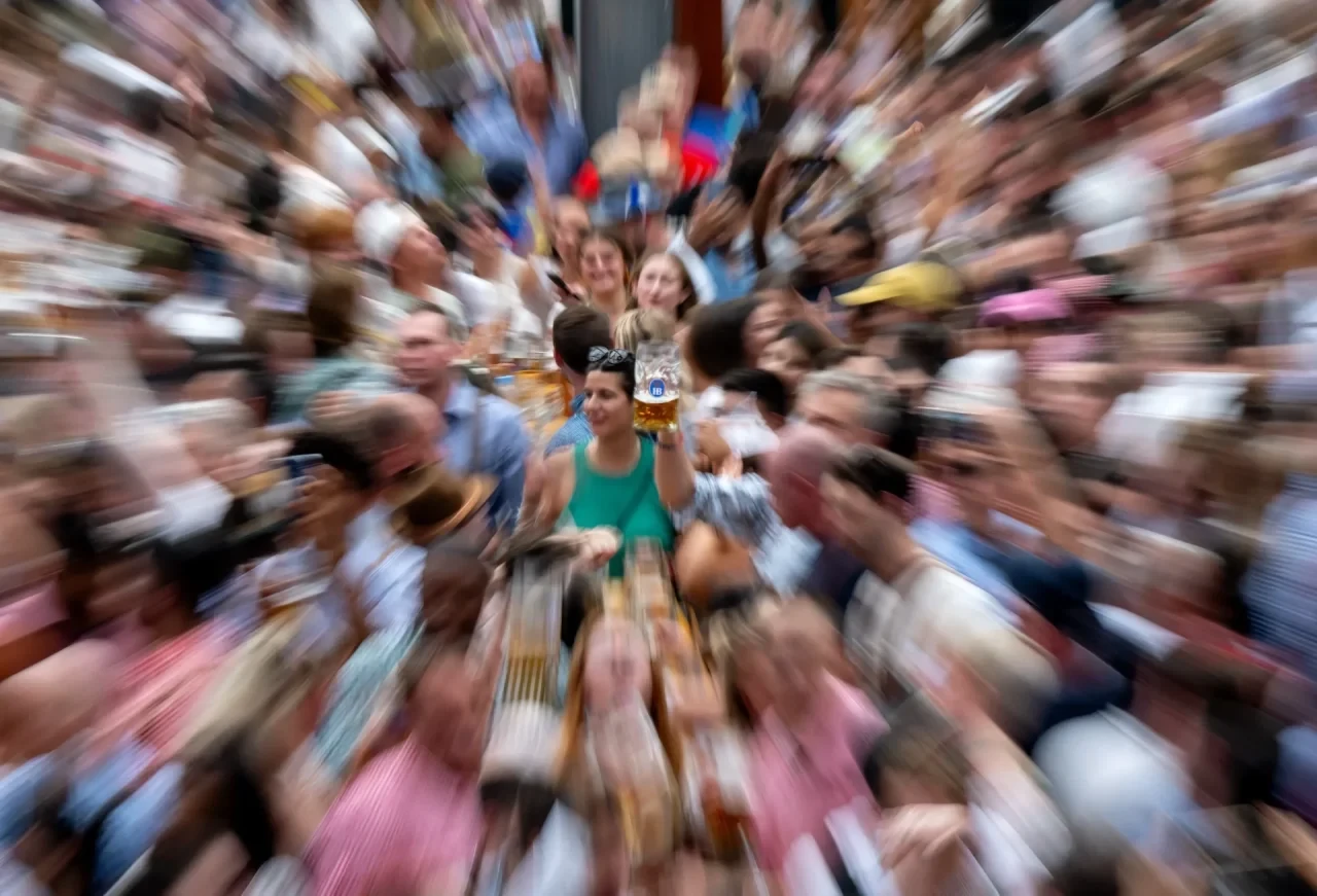 Unscharfe Aufnahme einer großen Menschenmenge in einem dicht besuchten Festzelt auf dem Oktoberfest. Im Bildzentrum sind mehrere Personen scharf erkennbar, während die Umgebung durch einen Zoom- bzw. Bewegungsunschärfe-Effekt verwischt dargestellt ist.