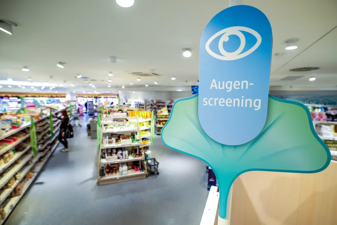 Augenscreening-Aufsteller mit stilisiertem Auge in einer dm-Filiale zwischen Verkaufsregalen