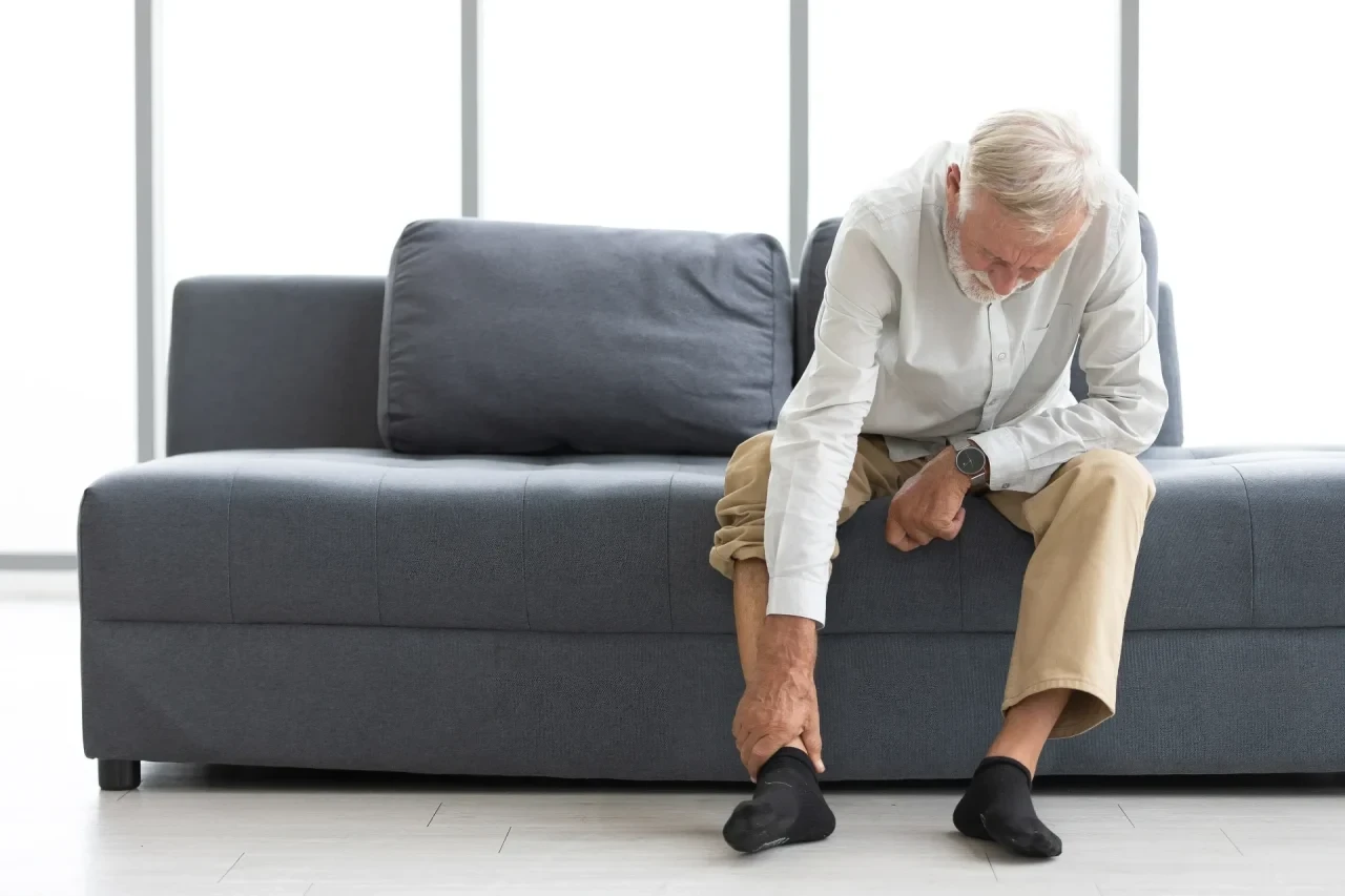 Älterer Mann sitzt auf einem Sofa und greift sich an den schmerzenden Fuß.