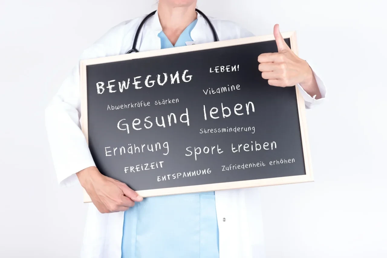Ärztin im weißen Kittel mit Stethoskop hält eine Tafel mit Begriffen zur Krebsprävention und einem gesunden Lebensstil – darunter Bewegung, Ernährung, Sport, Stressminderung und Entspannung – und zeigt den Daumen nach oben