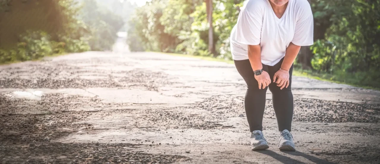 Person in Sportkleidung macht auf einer asphaltierten Straße im Grünen eine kurze Pause während des Trainings und stützt die Hände auf die Knie. Im Hintergrund sind Bäume und Sonnenlicht zu sehen.