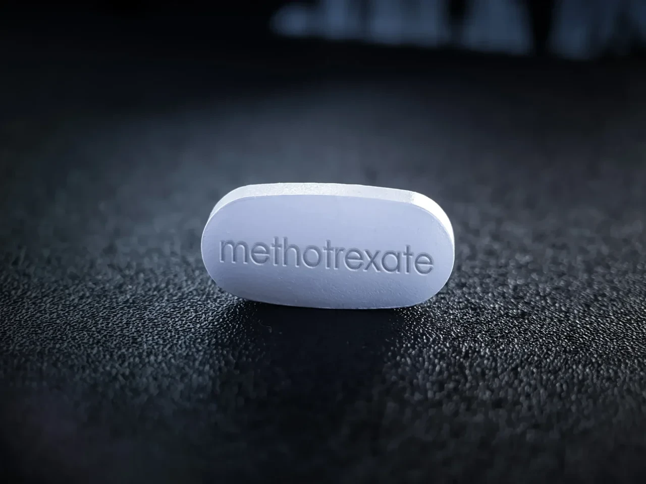 Weiße Methotrexat-Tablette mit der Prägung „methotrexate“ auf dunkler Oberfläche, aufgenommen in Nahaufnahme vor unscharfem Hintergrund.