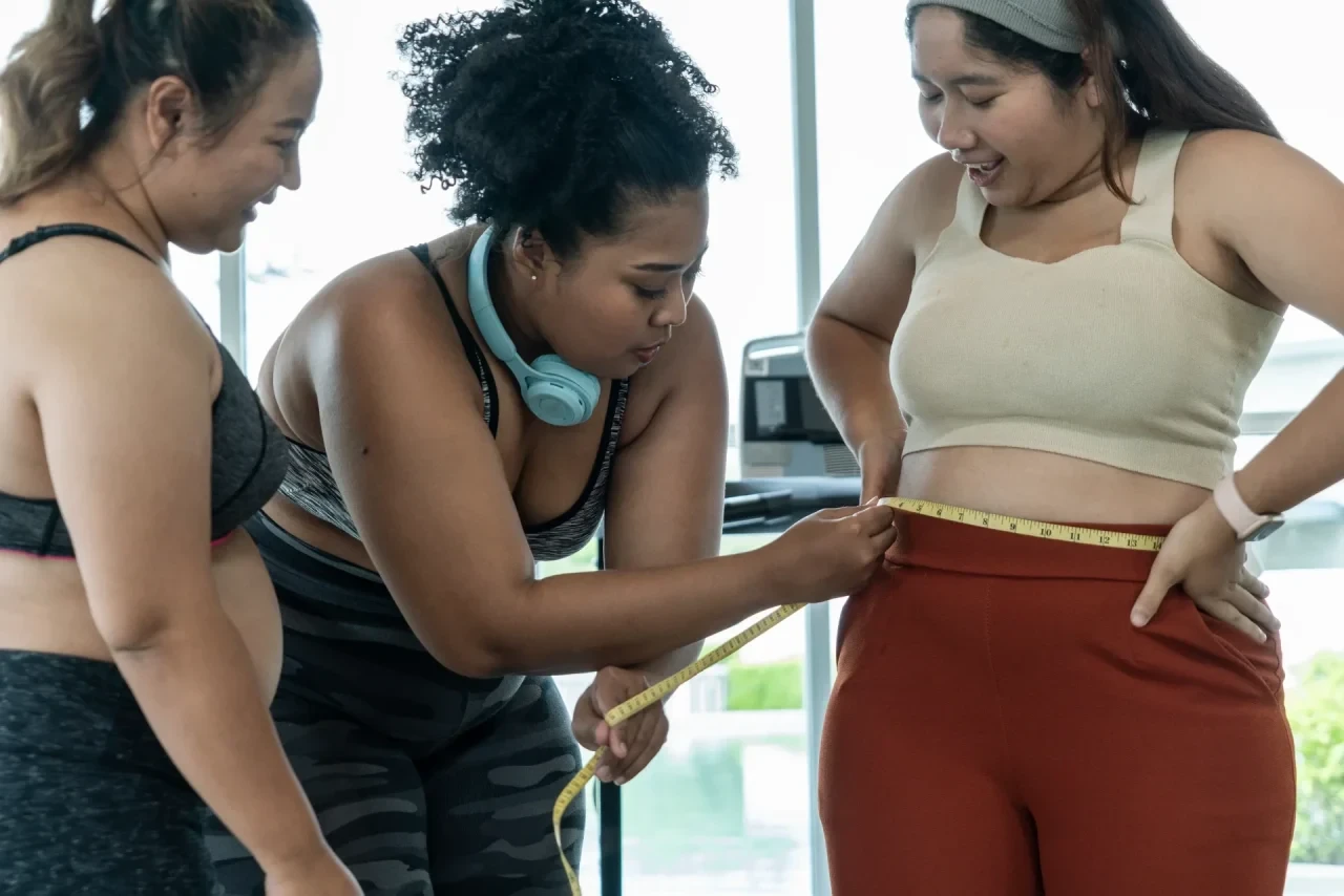Drei Personen unterschiedlicher Ethnien in Sportkleidung stehen in einem Fitnessstudio. Eine Person misst mit einem Maßband den Taillenumfang einer anderen Person. Im Hintergrund sind große Fenster und Fitnessgeräte zu sehen.