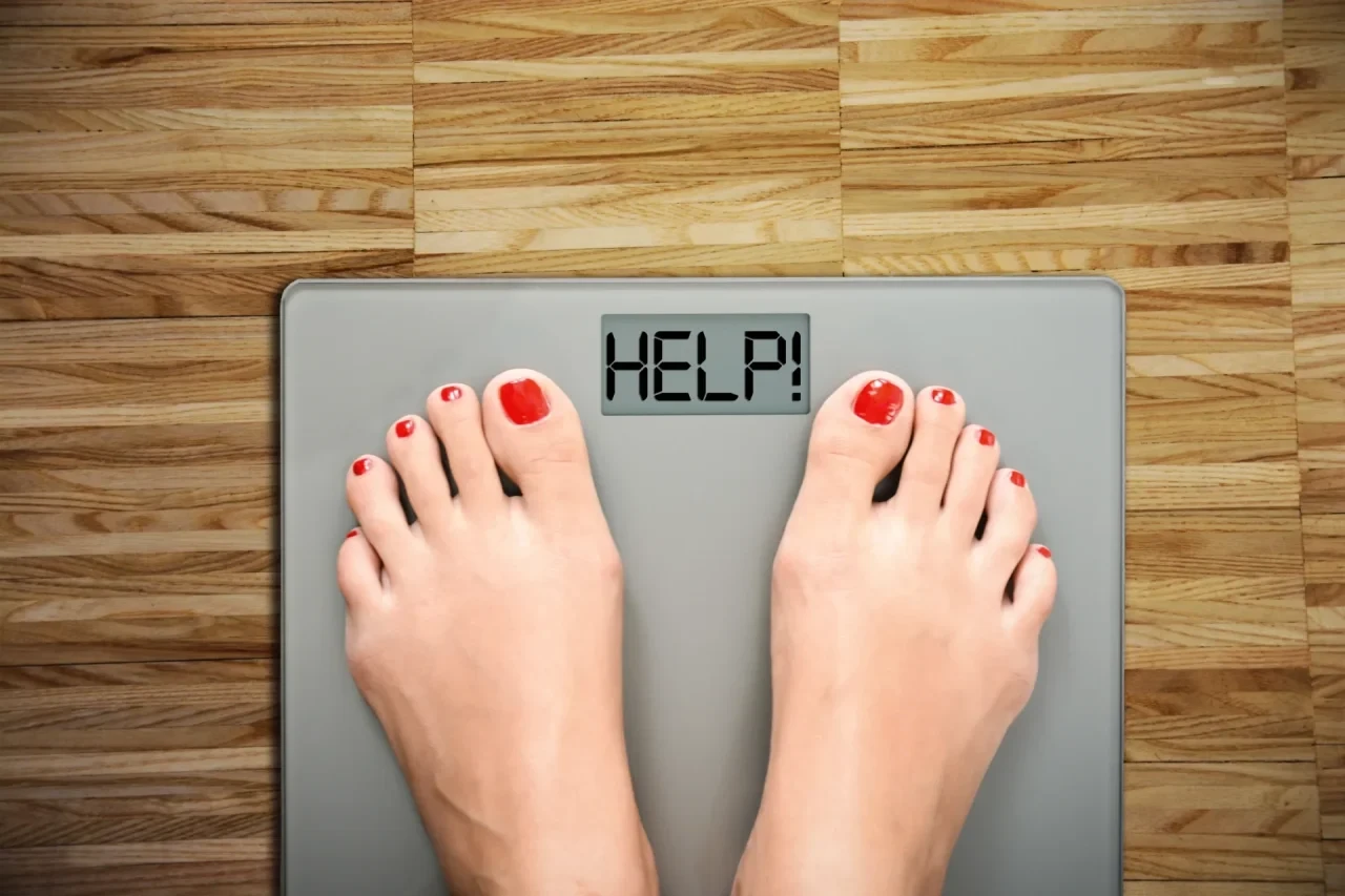 Füße einer Person auf einer digitalen Waage mit der Anzeige „HELP!“ vor einem Holzfußboden.