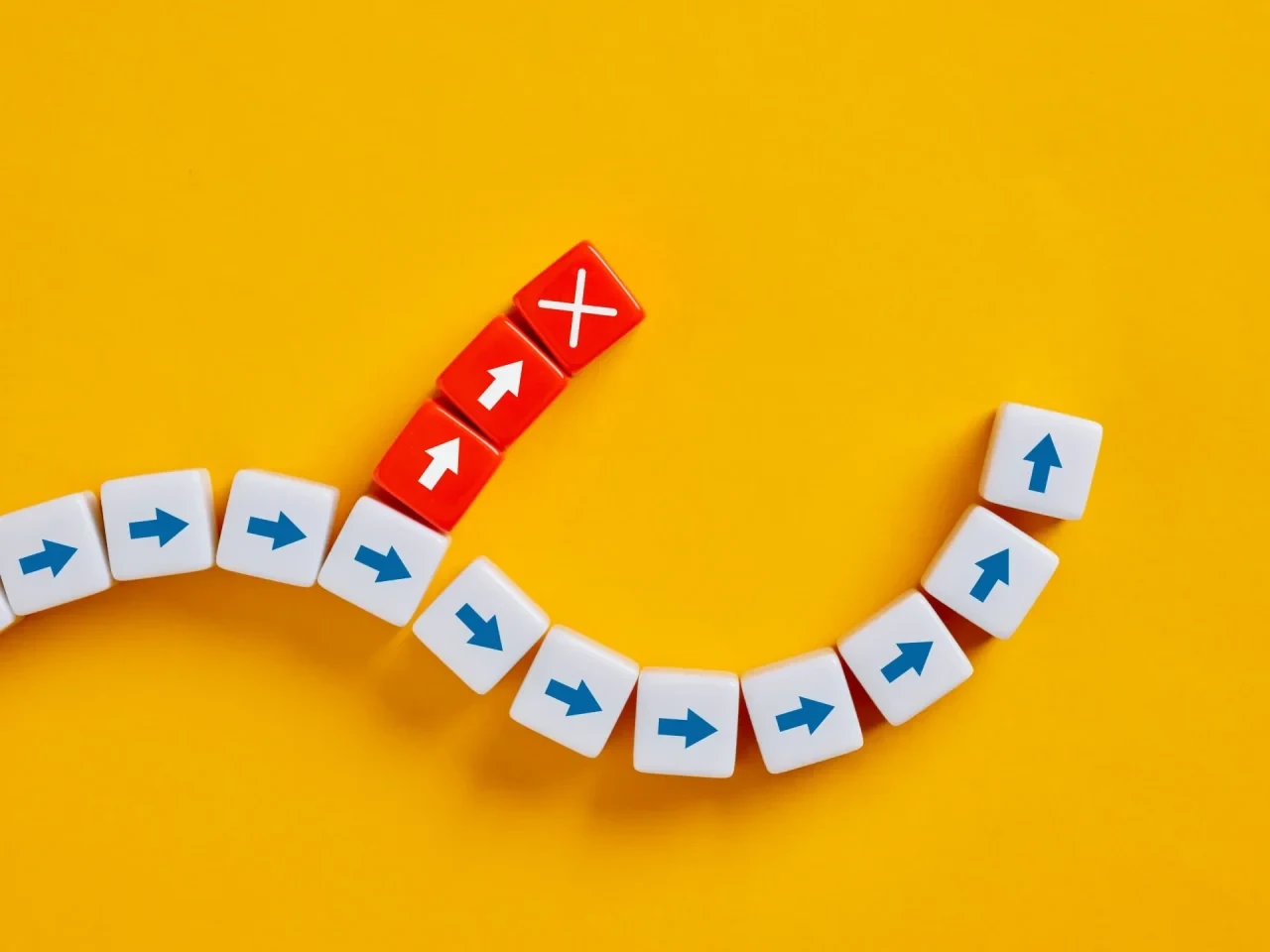 Weiße Spielsteine mit blauen Pfeilen bilden auf gelbem Hintergrund einen geschwungenen Weg. Ein abzweigender Pfad aus roten Spielsteinen endet in einem roten Stein mit weißem Kreuz und symbolisiert das gezielte Blockieren eines Signalwegs.