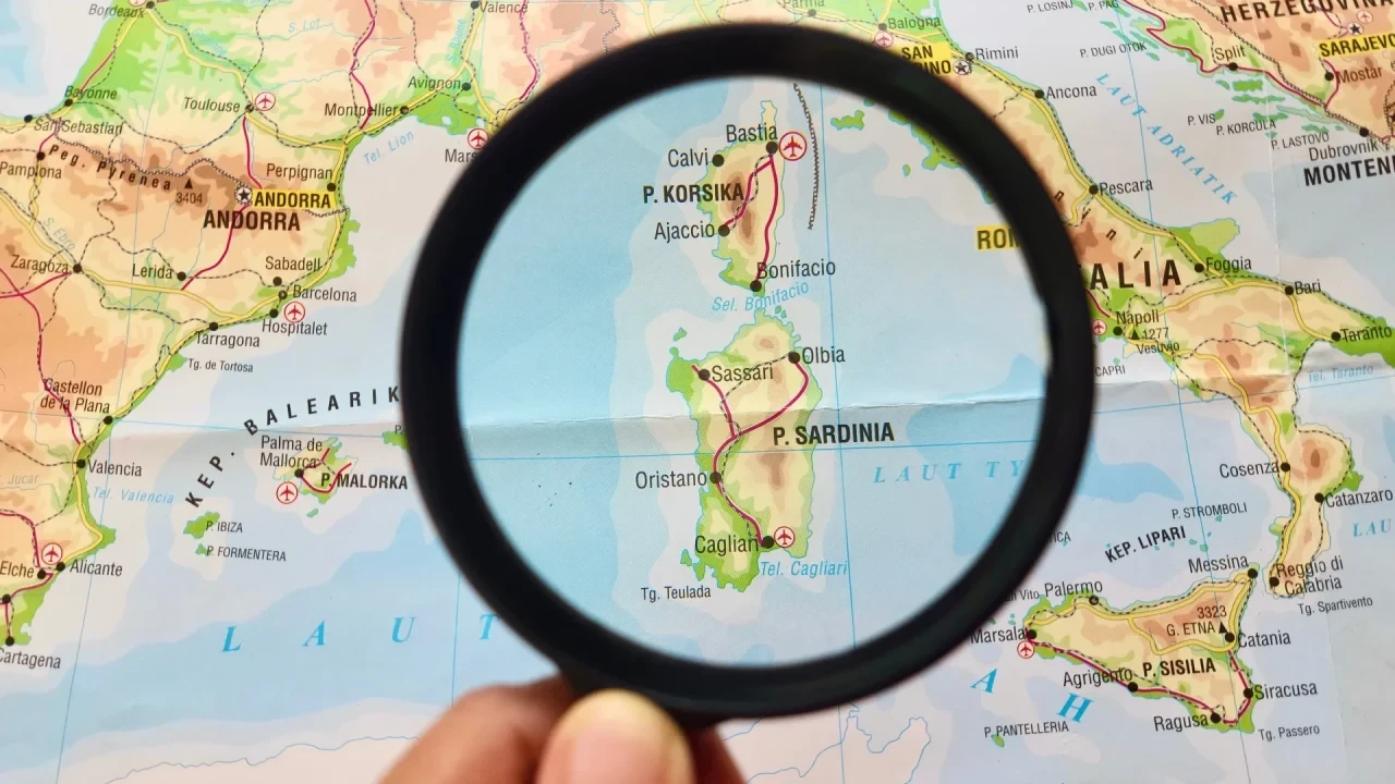 Landkarte des westlichen Mittelmeerraums mit Sardinien im Zentrum einer Lupe; sichtbar sind Teile Italiens, Korsikas, Spaniens und Frankreichs.