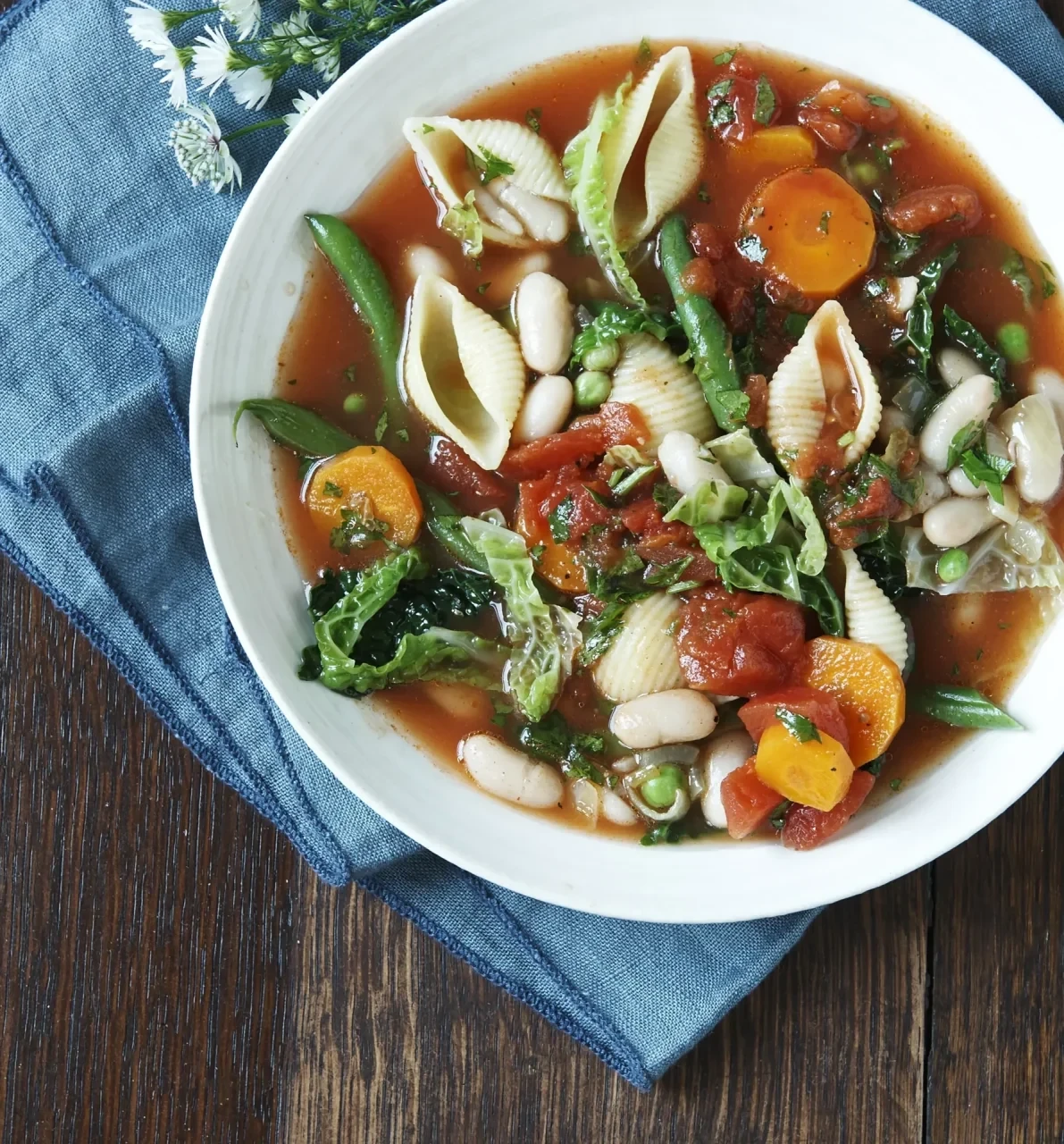 Gemüsereiche Suppe mit Karotten, Bohnen, Kohl, Tomaten und Nudeln in einer weißen Schüssel; Aufnahme aus der Vogelperspektive auf dunkler Holzoberfläche.