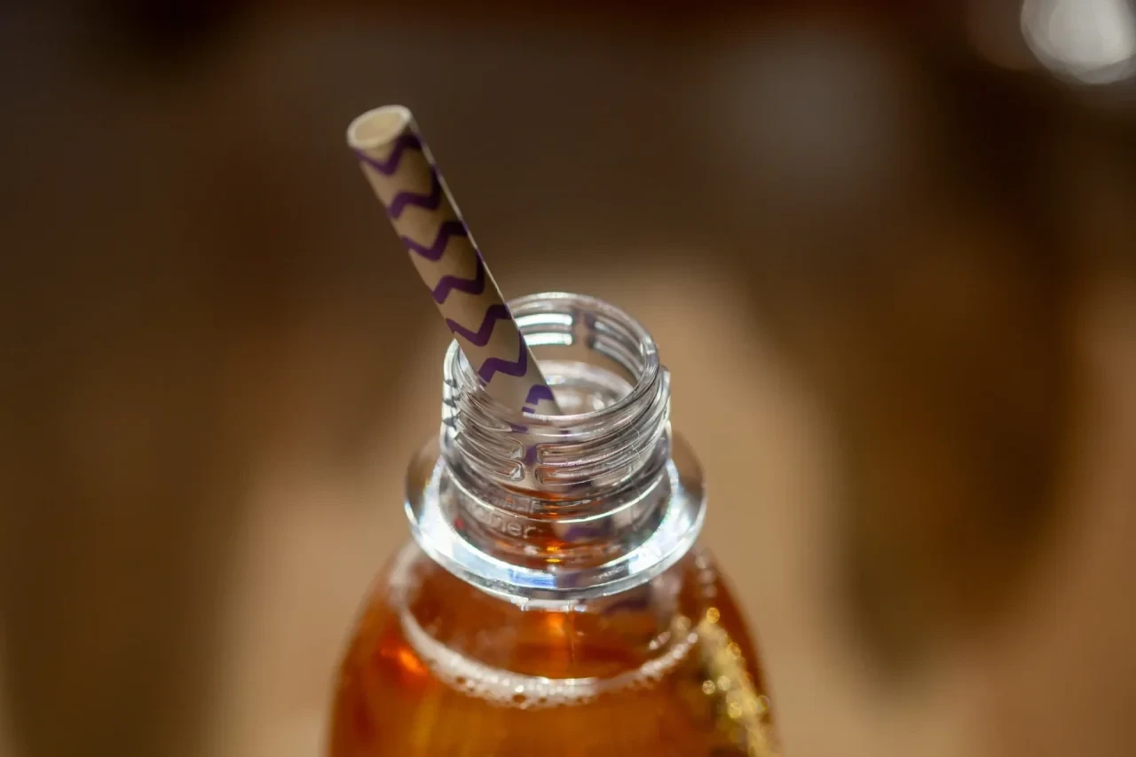 Eine geöffnete Flasche mit einem zuckerhaltigen Erfrischungsgetränk steht auf einem Tisch, in der Öffnung steckt ein Trinkhalm.