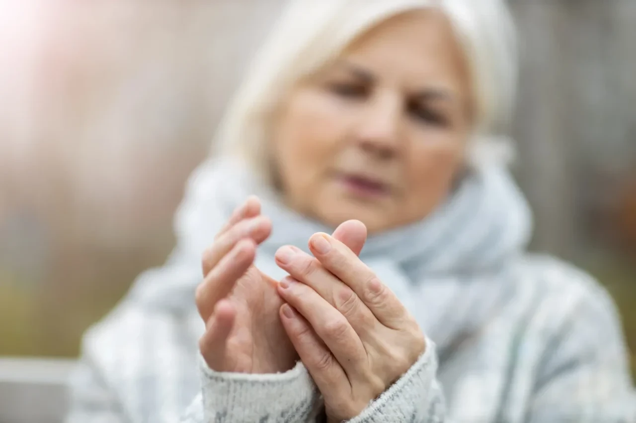 Eine ältere Frau in heller Kleidung hält eine Hand mit der anderen und betrachtet die Finger. Die Hände sind scharf fokussiert, während der Hintergrund und das Gesicht unscharf erscheinen.