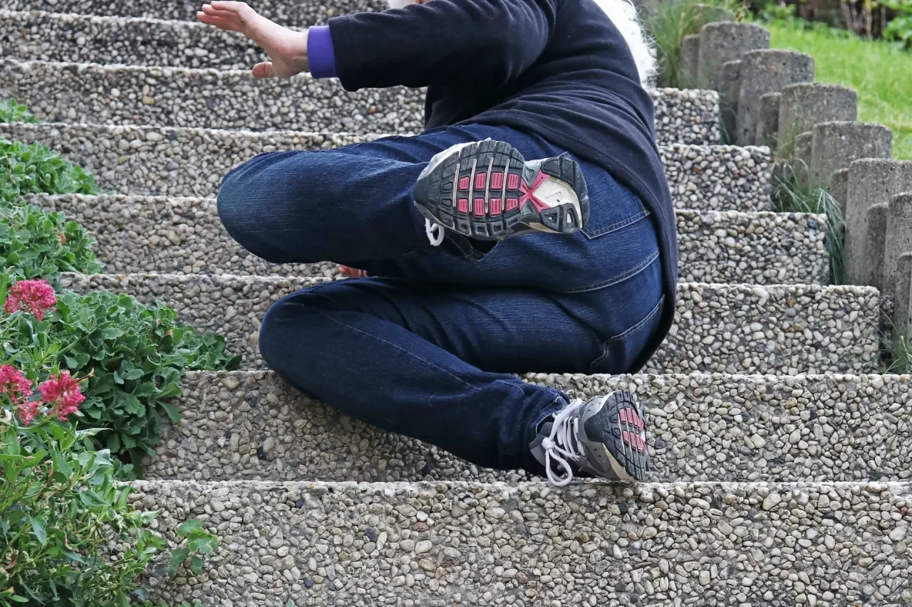 Eine Frau liegt nach einem Sturz auf einer Außentreppe aus Betonstufen; ein Bein ist angewinkelt, ein Arm ausgestreckt.