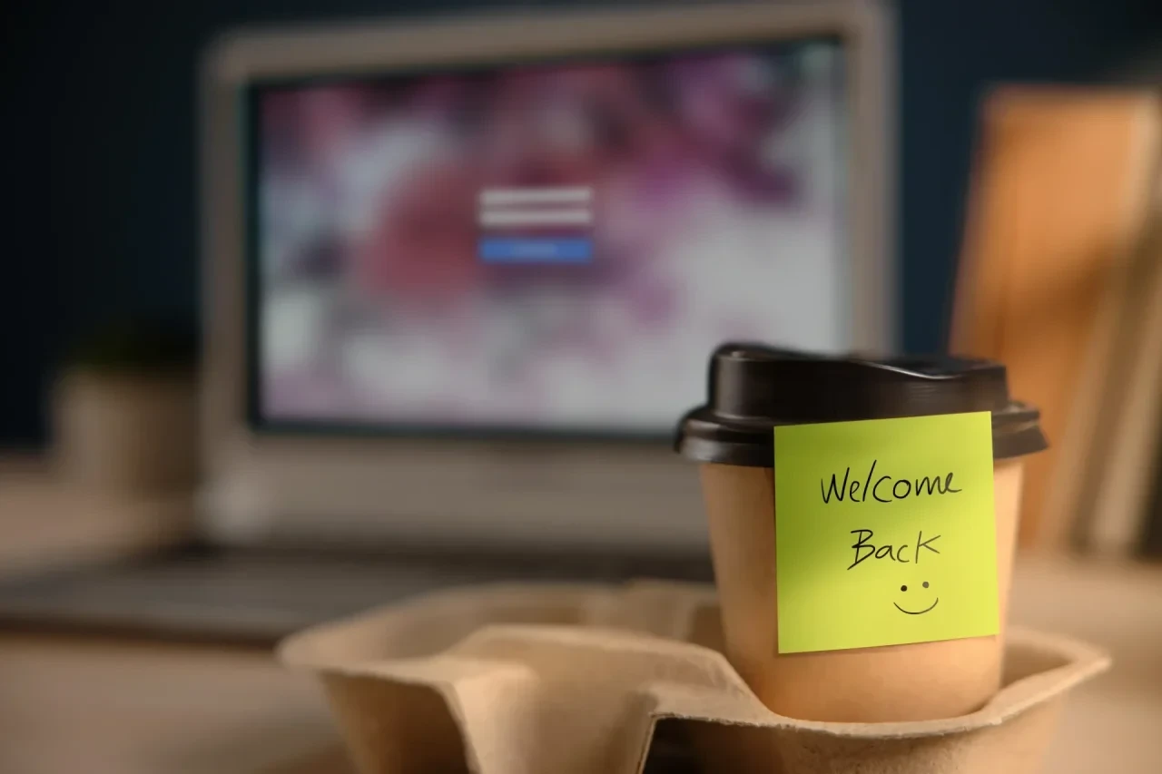 Kaffeebecher mit einem gelben Haftzettel mit der Aufschrift „Welcome Back“ vor einem unscharf dargestellten Laptop-Arbeitsplatz.