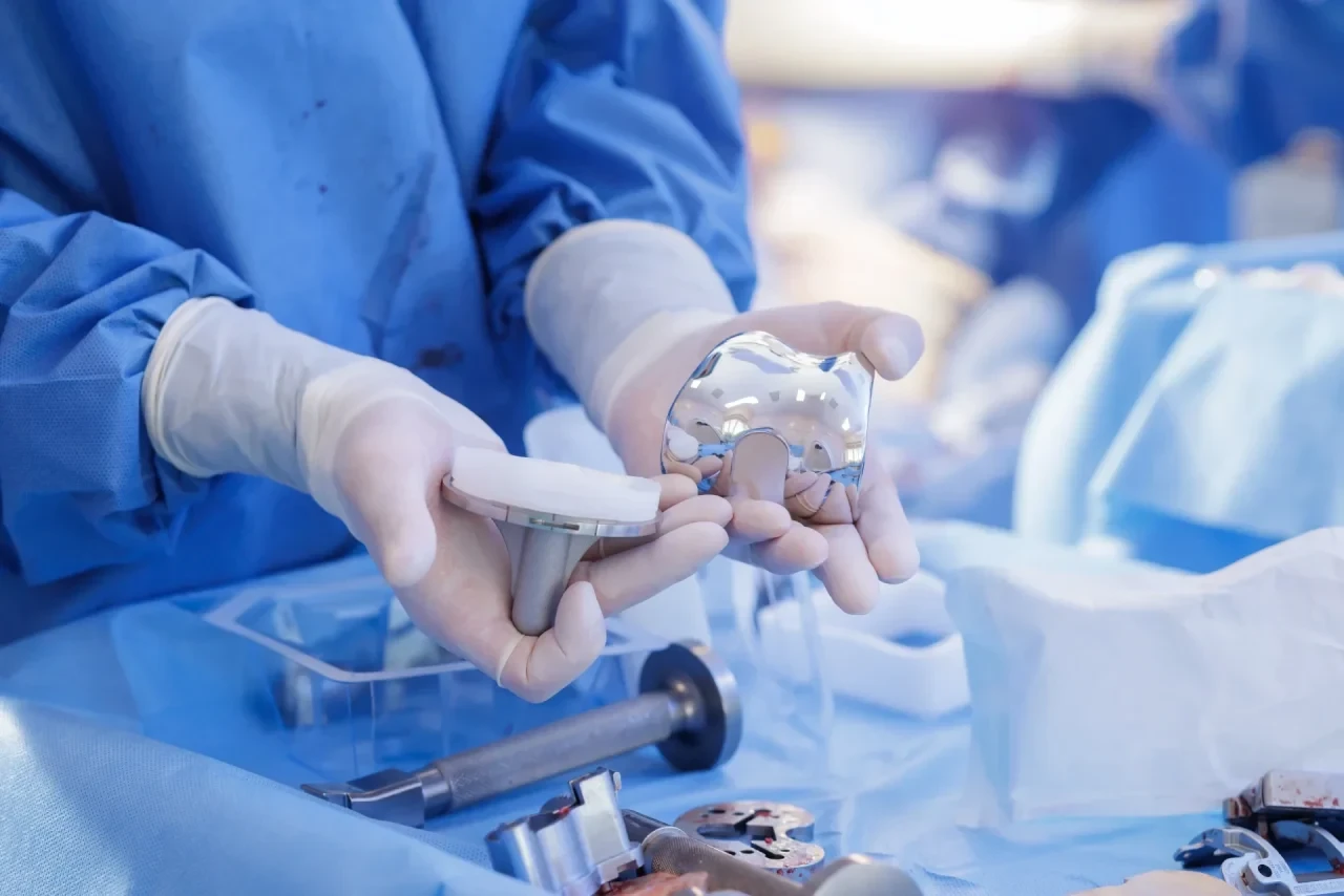 Behandschuhte Hände halten die metallischen und kunststoffbasierten Komponenten einer Kniegelenksprothese in einem Operationssaal; im Hintergrund sind weitere sterile Materialien und Instrumente zu sehen.
