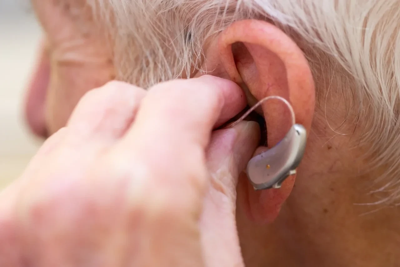 Detailaufnahme eines Ohrs einer älteren Person mit einem Hinter-dem-Ohr-Hörgerät. Zu sehen ist eine Hand, die das Gerät gerade einsetzt.