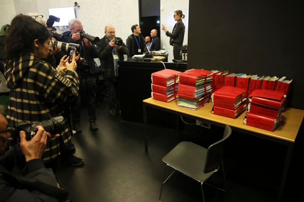 Fotografen, Angeklagte und Justizbeamte vor Schreibtisch mit vielen roten Akten.