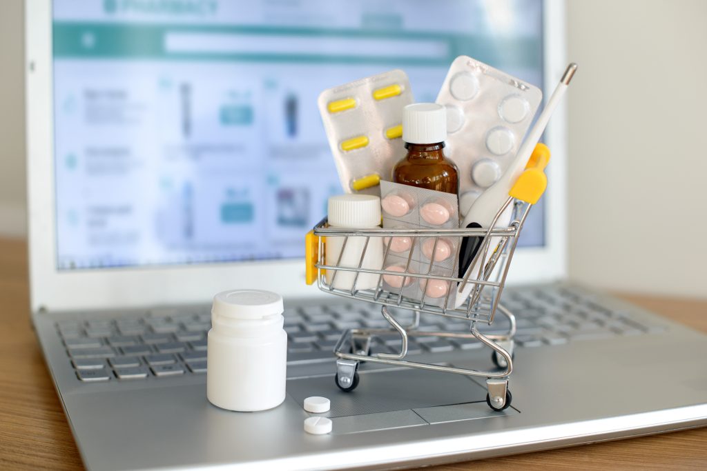 Einkaufswagen mit Medikamenten vor einem Laptop-Bildschirm mit der Website einer Apotheke. Pillen, Blisterpackungen, medizinische Flaschen, Thermometer-Set. Gesundheitswesen und Online-Shopping.