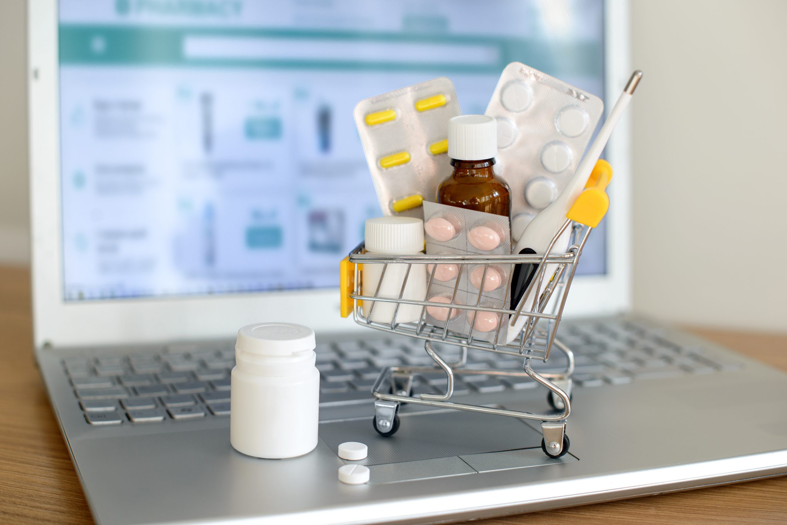 Einkaufswagen mit Medikamenten vor einem Laptop-Bildschirm mit der Website einer Apotheke. Pillen, Blisterpackungen, medizinische Flaschen, Thermometer-Set. Gesundheitswesen und Online-Shopping.